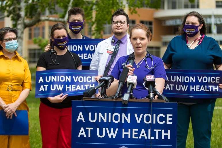 A person in scrubs holds a microphone as people behind the person hold signs reading “A UNION VOICE AT UW HEALTH.”