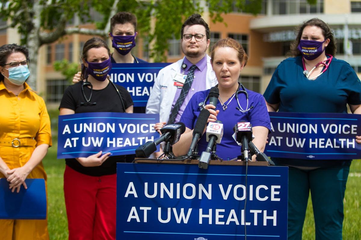 A person in scrubs holds a microphone as people behind the person hold signs reading “A UNION VOICE AT UW HEALTH.”