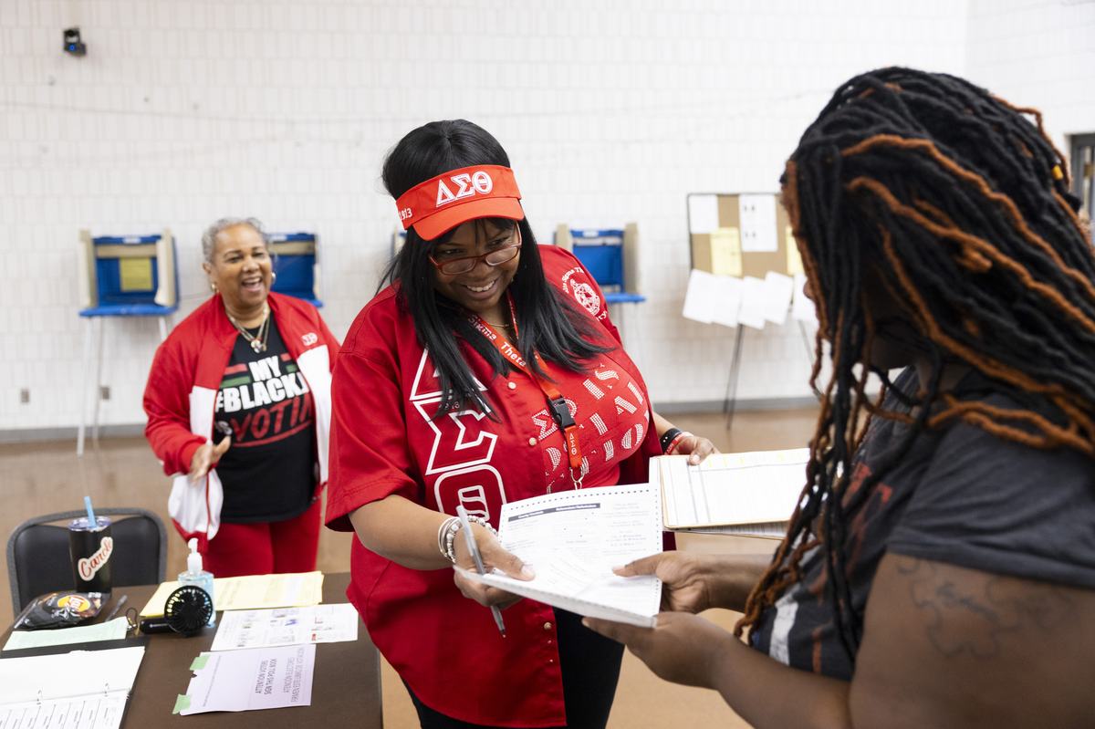 A person in a red shirt and visor hands a form to another person next to a table covered with papers and other items, with another person, voting booths and a bulletin board visible.