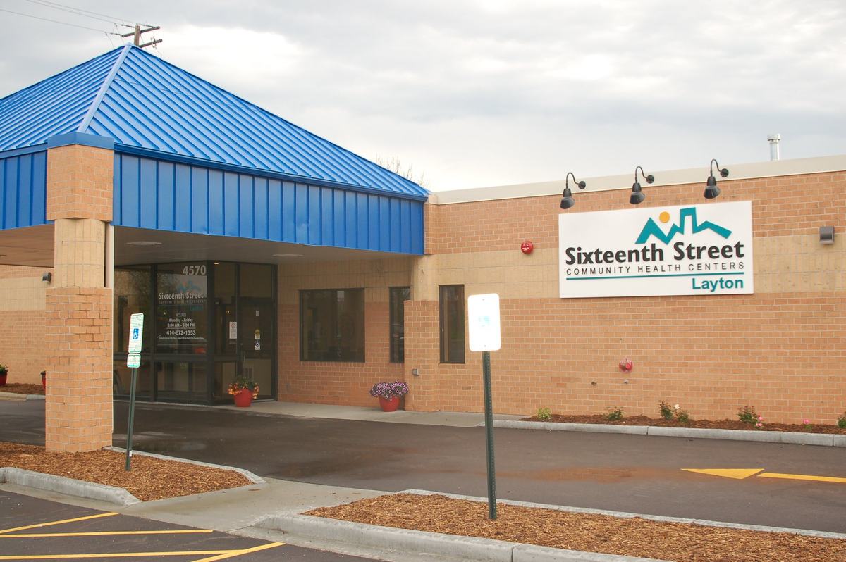 A brick building with a blue metal roof over the entrance displays a sign reading "Sixteenth Street Community Health Centers Layton"