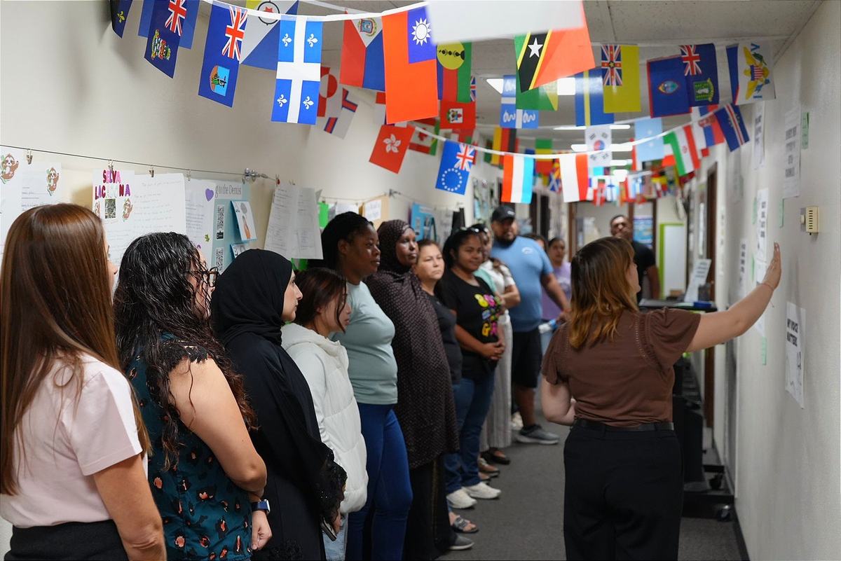 People stand in a line in a hallway as a person points to papers on a wall beneath rows of hanging international flags.