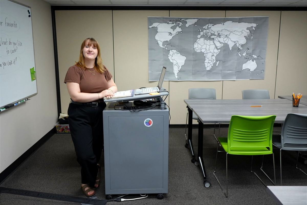 A person stands beside a laptop on a lectern in a room with a world map, a whiteboard, tables and green and gray chairs.