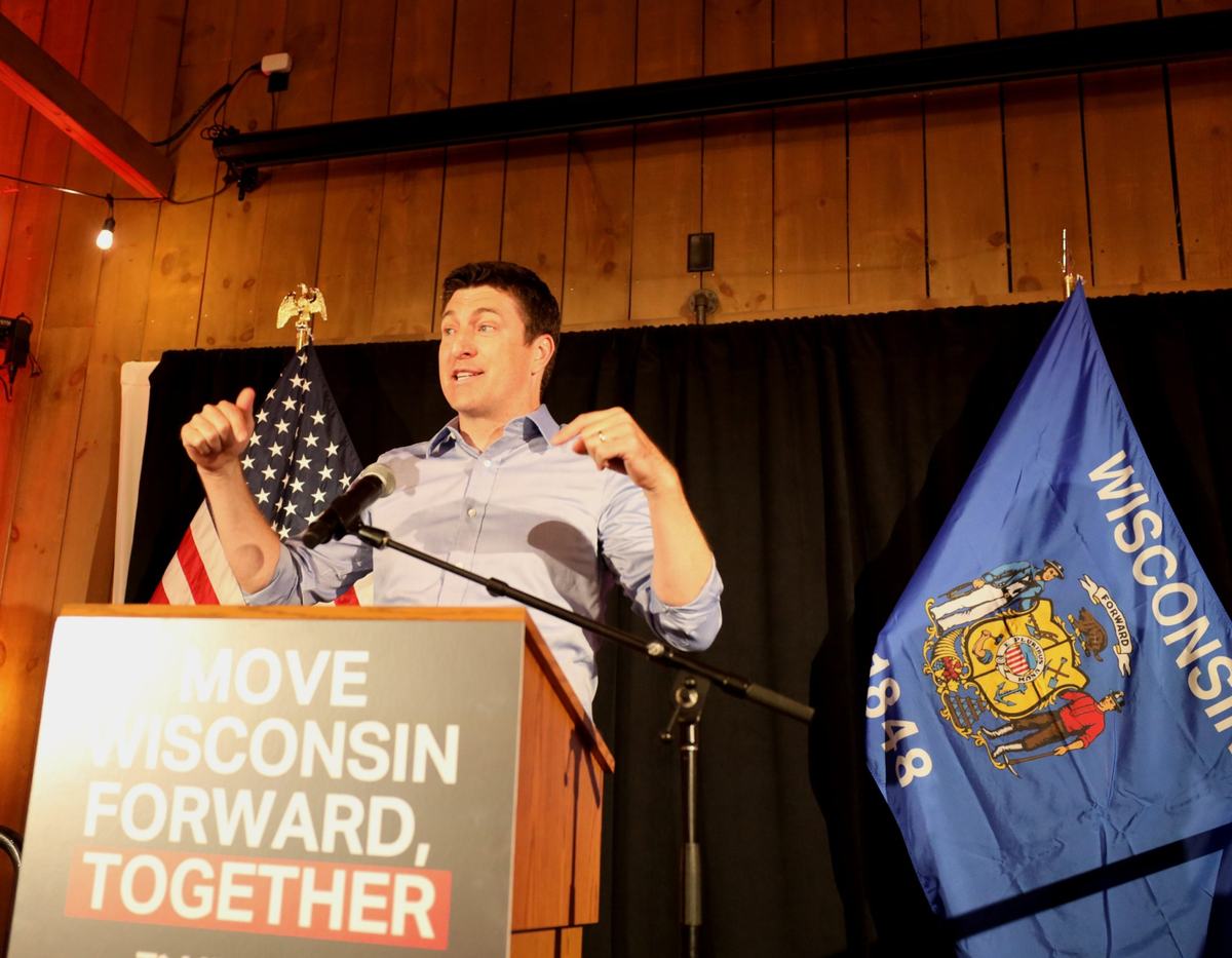 A person in a light blue shirt gestures with both hands while standing at a podium reading "Move Wisconsin Forward, Together," flanked by U.S. and Wisconsin flags.