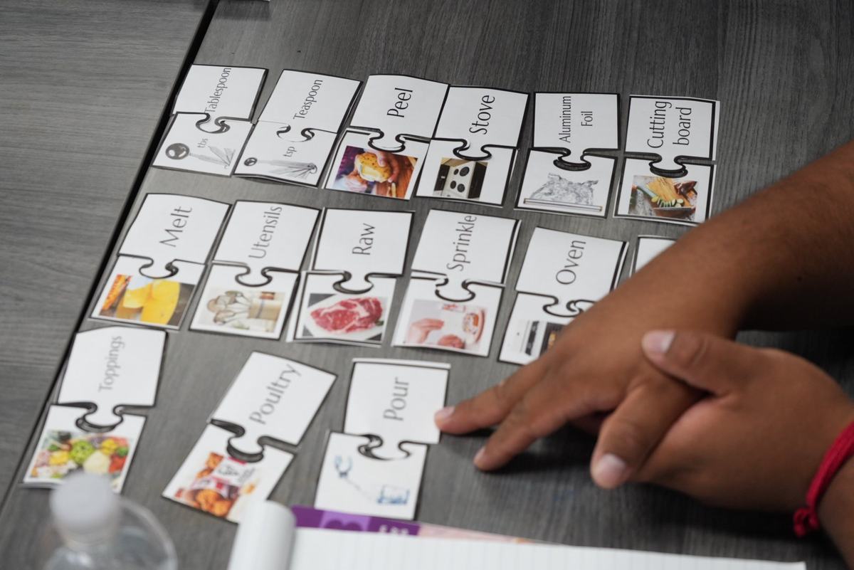Hands rest beside matching puzzle cards labeled with cooking terms including "Tablespoon," "Peel," "Stove," "Melt," "Raw" and "Oven"