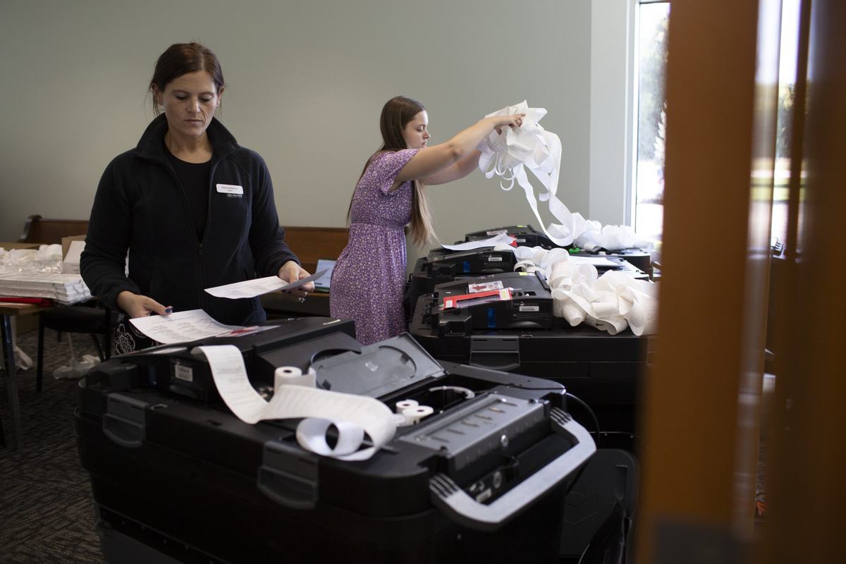 Two women work with voting machines during an election equipment test, examining paperwork and handling long strips of printed paper ballots.