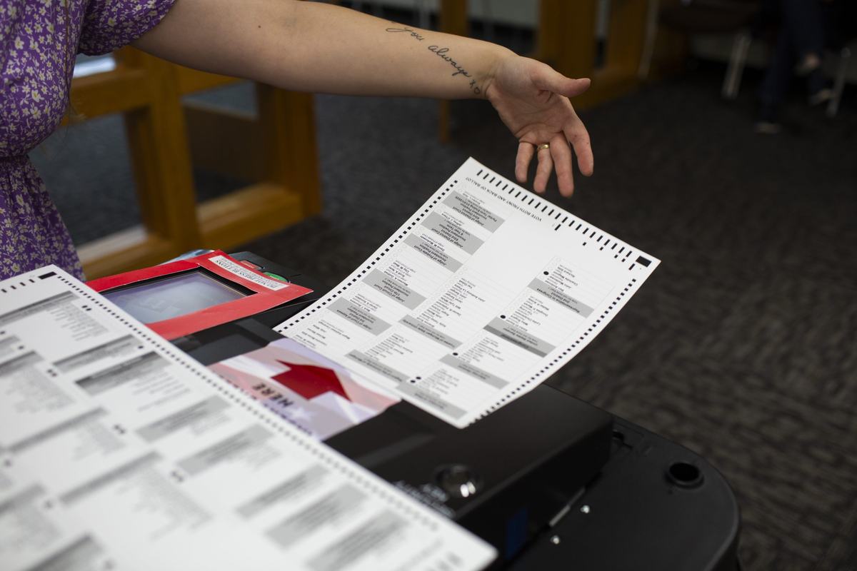 A person gestures toward a paper election ballot positioned above a voting machine during a demonstration of voting equipment.