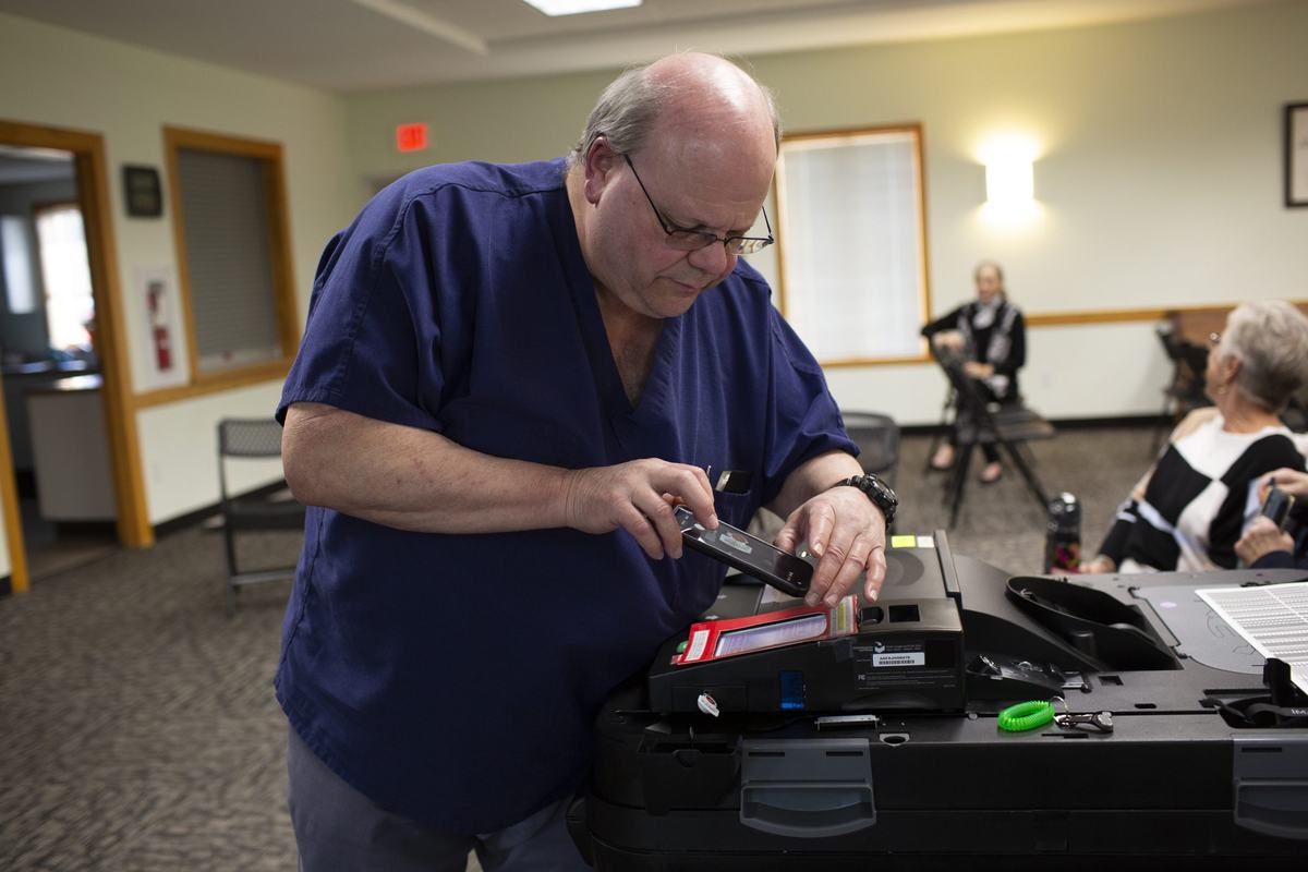 A man wearing navy blue scrubs and glasses examines voting equipment while holding a smartphone in a room with several seated people.