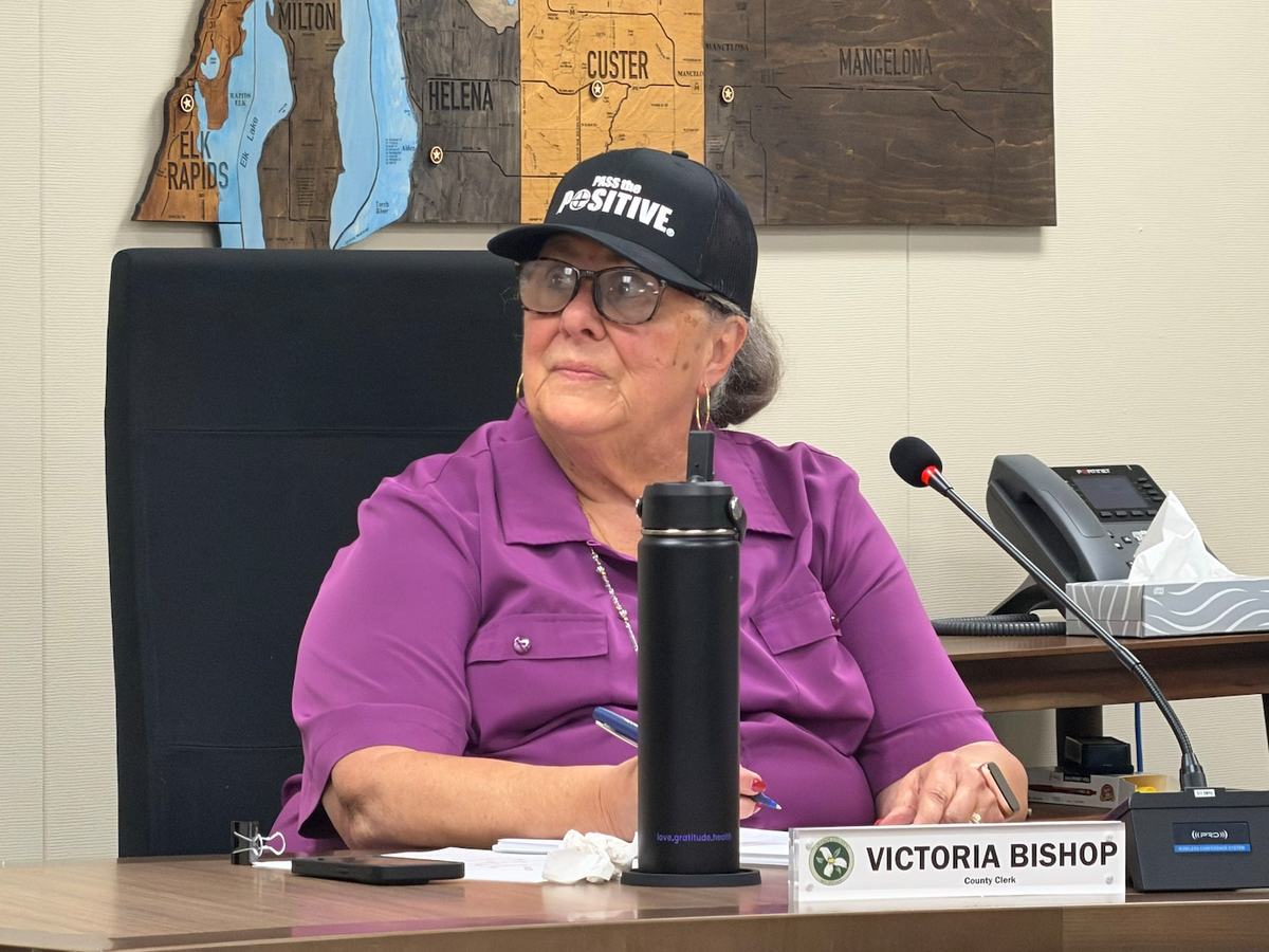 A white woman wearing a purple shirt and a black baseball cap sits at a table in a meeting room.