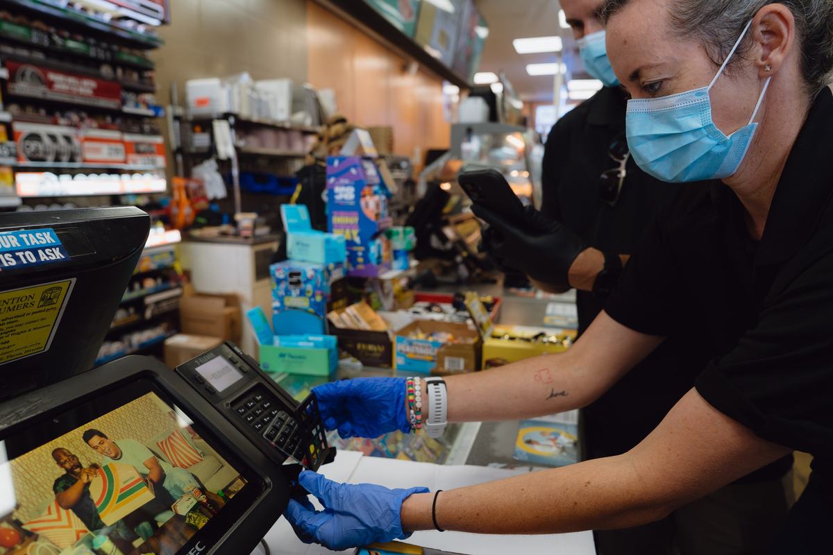 A person wearing blue gloves removes an overlay from a card reading machine at a convenience store checkout counter.