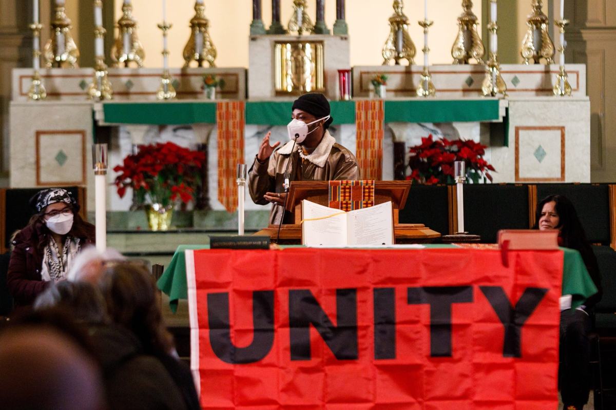 A person wearing a face mask stands and motions with hands at a lectern inside a church behind a large red banner reading "UNITY" with other people seated.