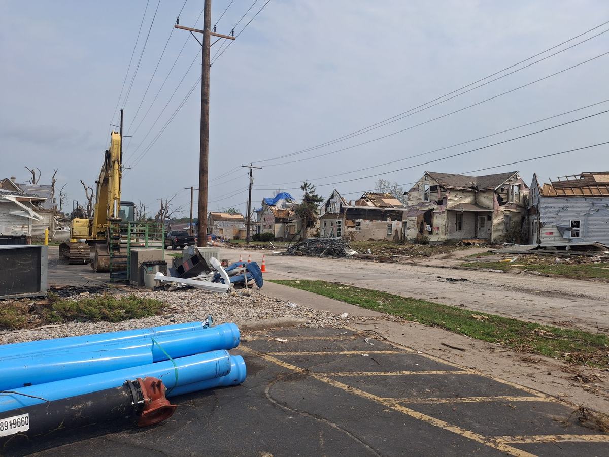 Damaged houses line a debris-strewn street with broken trees and utility poles as a yellow excavator and large blue pipes sit nearby.