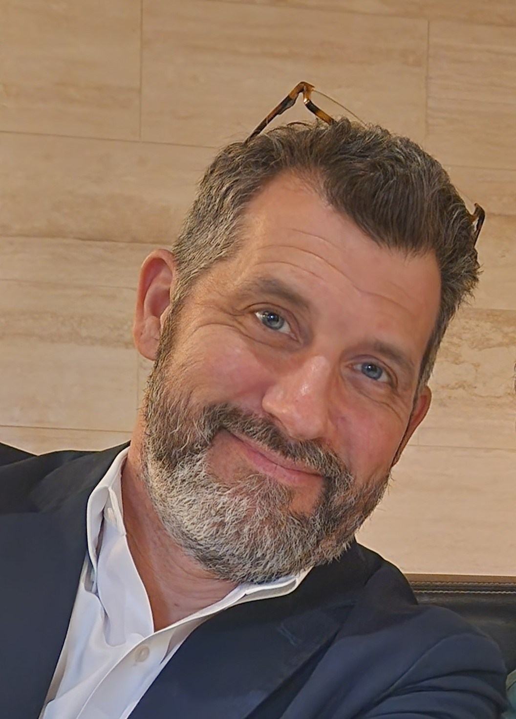 A person with short dark hair and a gray beard wears a dark jacket over a white collared shirt, with glasses resting on the person's head.