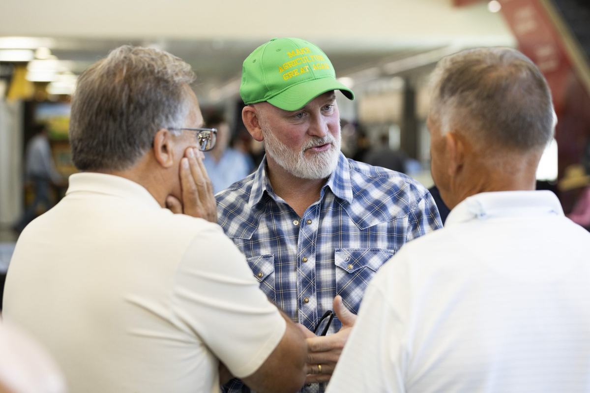 A person wearing a green hat reading "Make Agriculture Great Again" and a blue plaid shirt stands facing two people in white shirts.