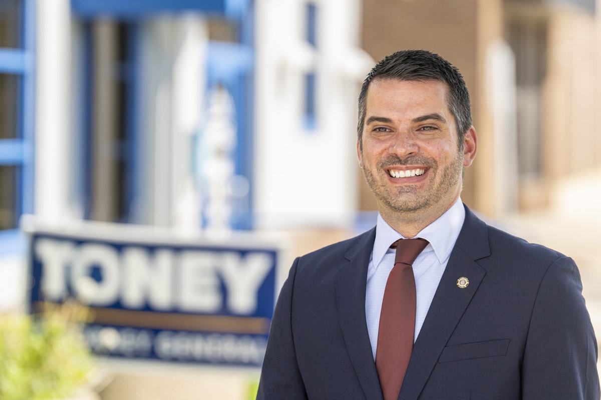 A person in a dark suit and red tie stands near a blurred sign reading "TONEY"