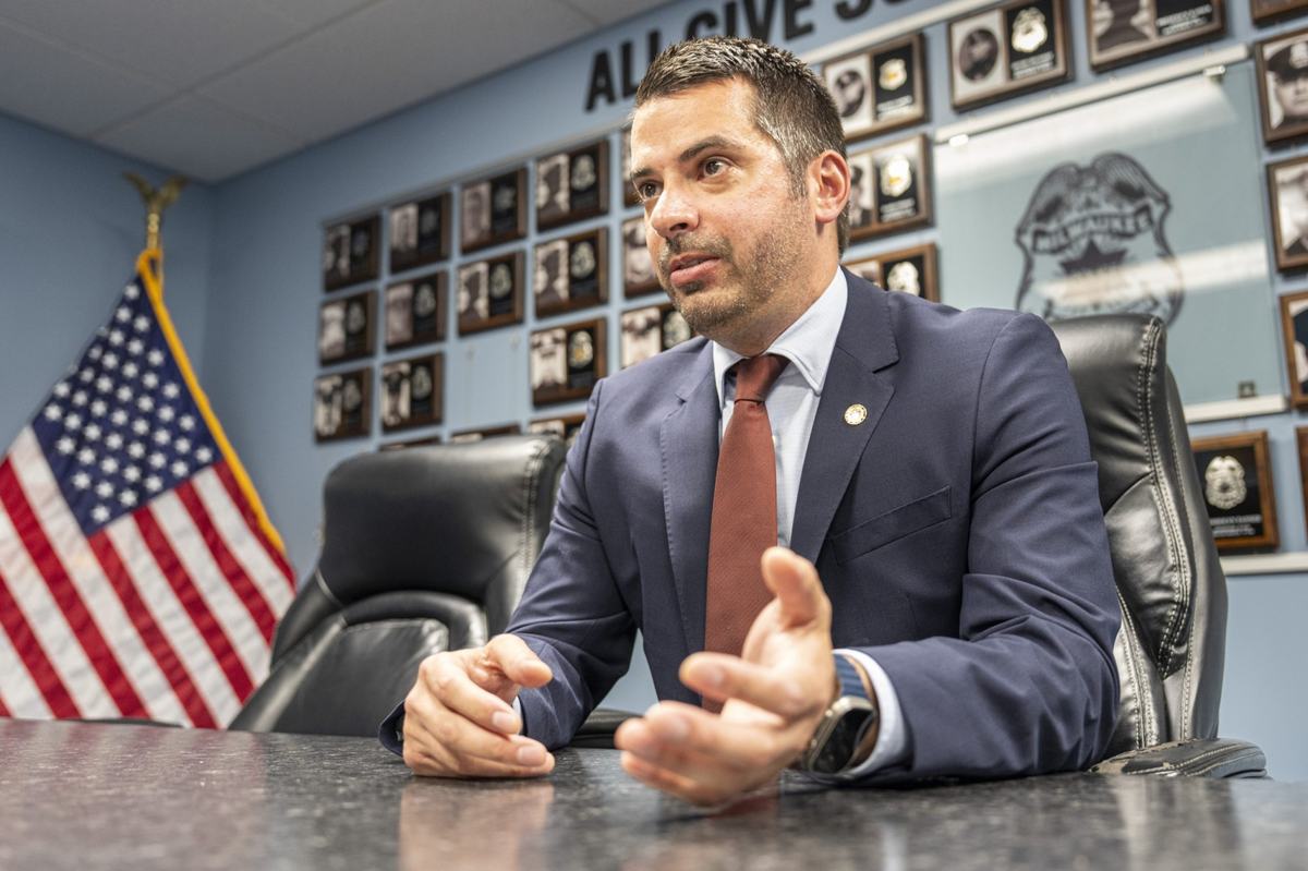 A person in a suit gestures while seated at a table, with an American flag and a wall of framed plaques.