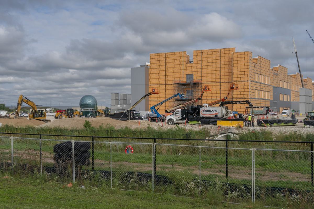 A large building with yellow exterior panels stands under construction, surrounded by excavators, boom lifts, trucks and people in safety vests behind a fence.
