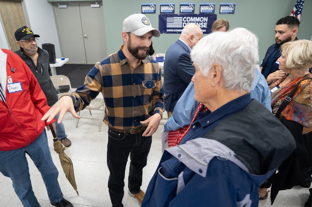 A person in a plaid shirt and gray cap gestures while standing with people in a room with "Berman" campaign signs and a "Common Defense" banner on a wall.