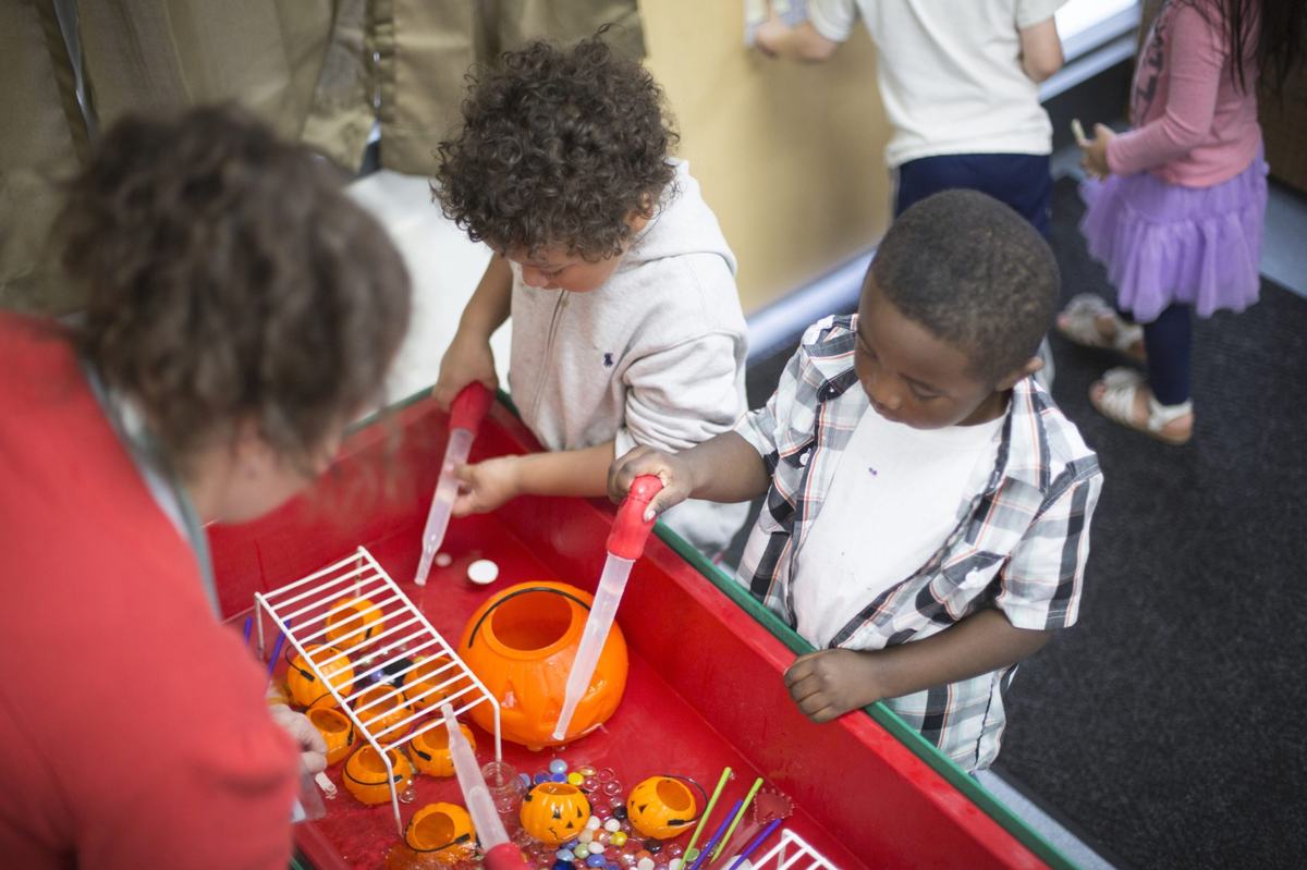Two people use large plastic droppers at a red activity table filled with orange pumpkin containers, colorful beads and water as others stand nearby.