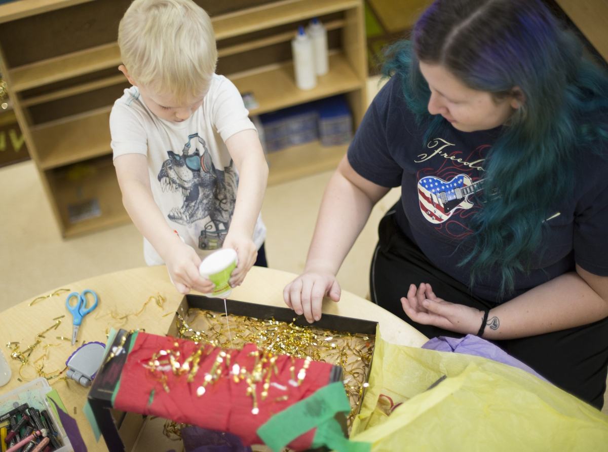 A person with blond hair squeezes glue onto gold tinsel inside a cardboard box while another person with blue and purple hair watches at a table covered with craft supplies.