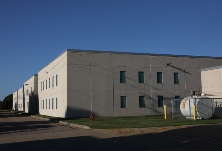 A large white building with gray along the top stands beside a paved drive, with a fenced metal tank and fire hydrant outside.