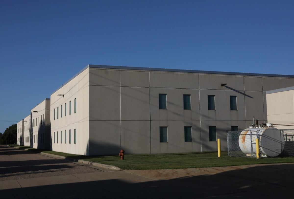 A large white building with gray along the top stands beside a paved drive, with a fenced metal tank and fire hydrant outside.