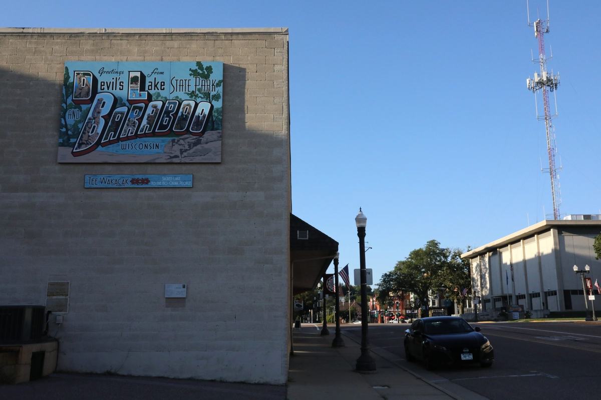 A street beside a building with a mural reading "Greetings From Devil's Lake State Park and Baraboo Wisconsin," with a communications tower nearby.
