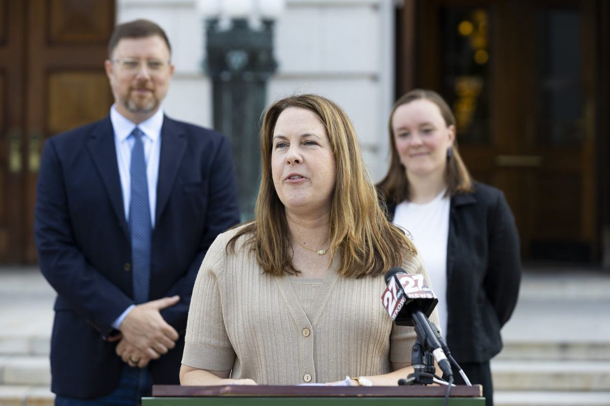 A person stands at a podium with a WKOW 27 News microphone while two people stand in the background outside a building.