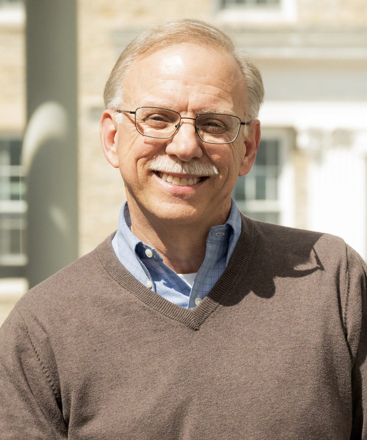 A person with glasses and a white mustache wears a brown V-neck sweater over a blue collared shirt outdoors with a building in the background.
