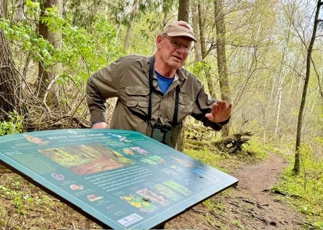 A person wearing a cap, a khaki shirt and binoculars gestures beside an informational sign along a wooded trail.