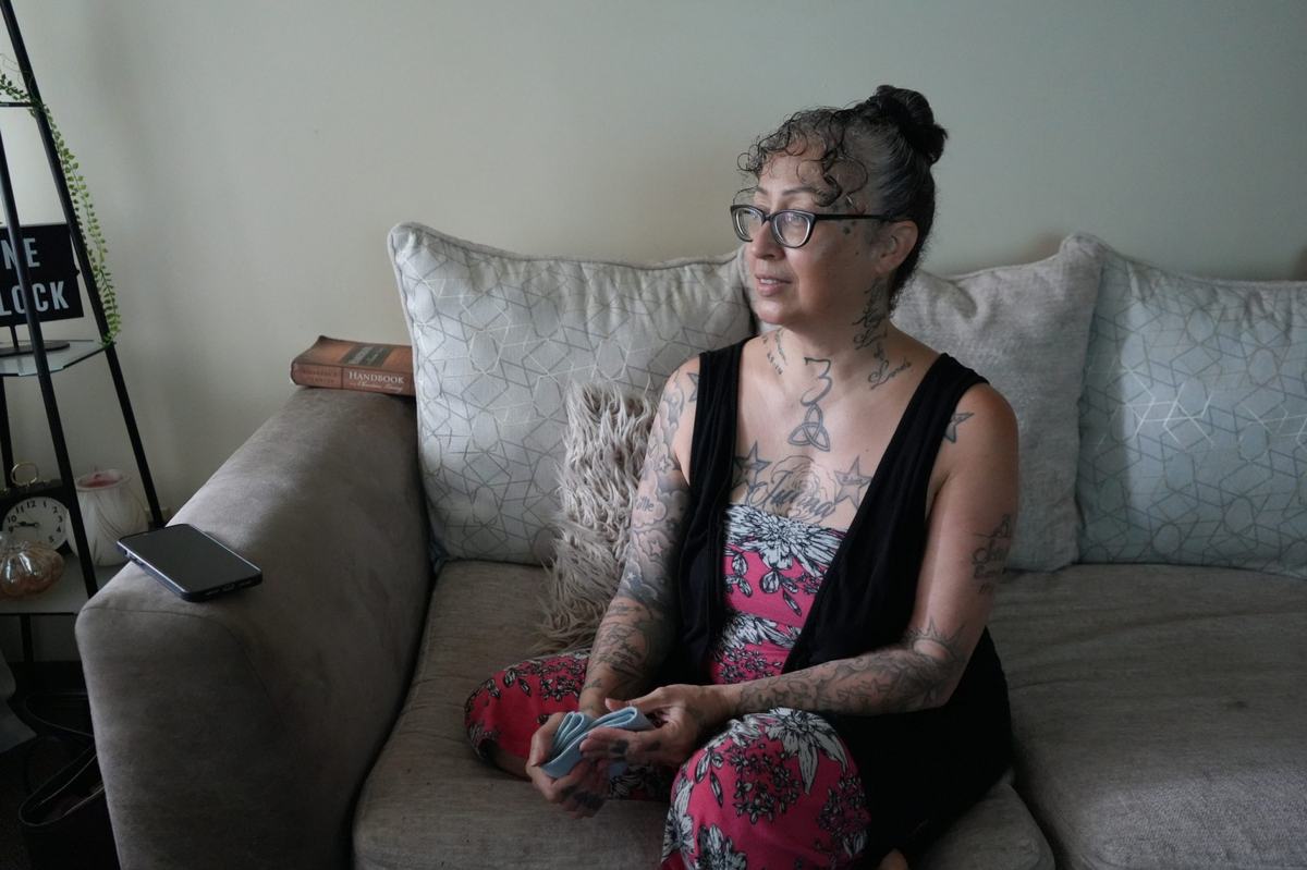 A person wearing glasses sits on a couch holding a folded cloth, with a cellphone, a book and other items nearby.