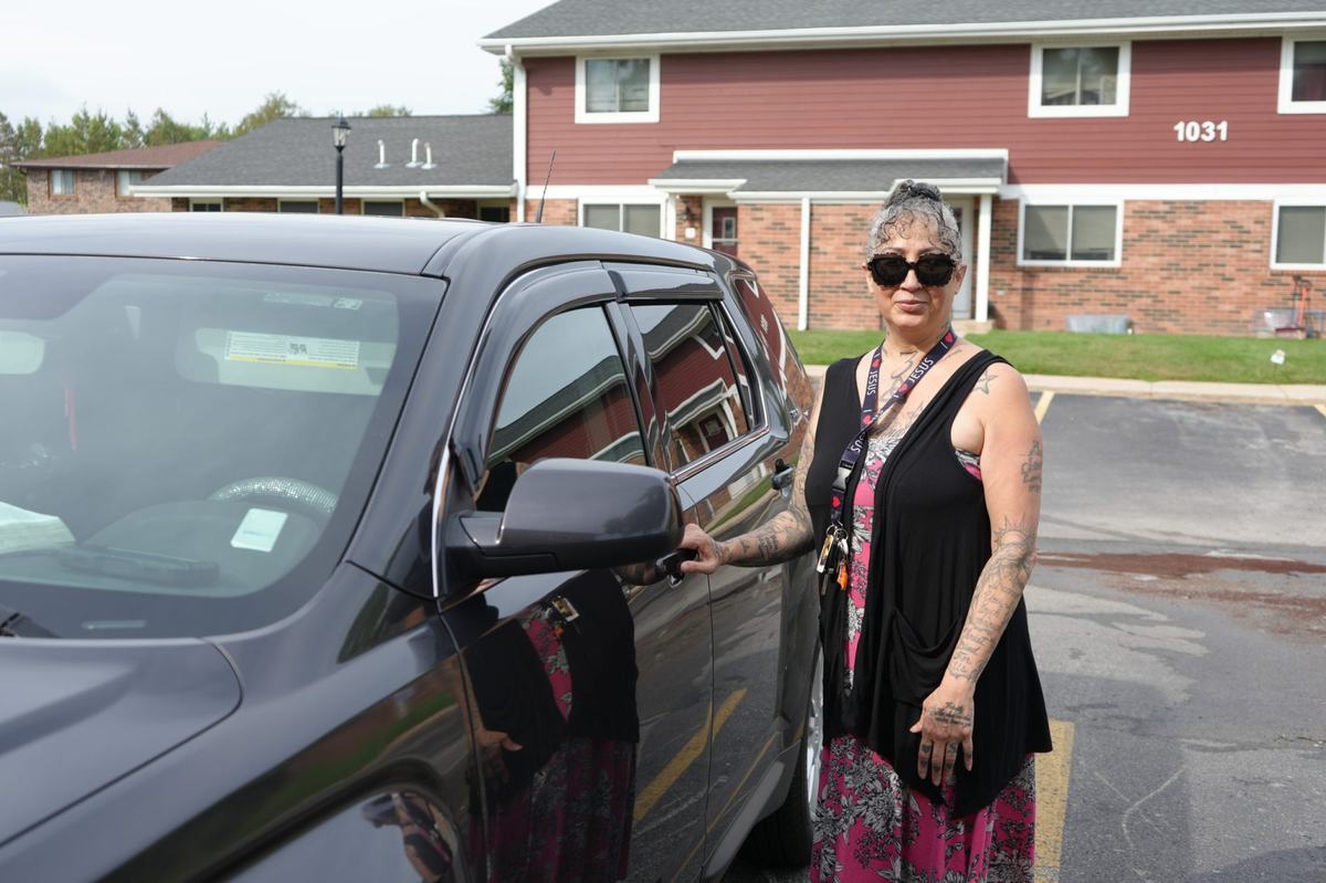 A person wearing sunglasses and a lanyard stands beside a dark vehicle in a parking lot, with a red and brick building in the background.