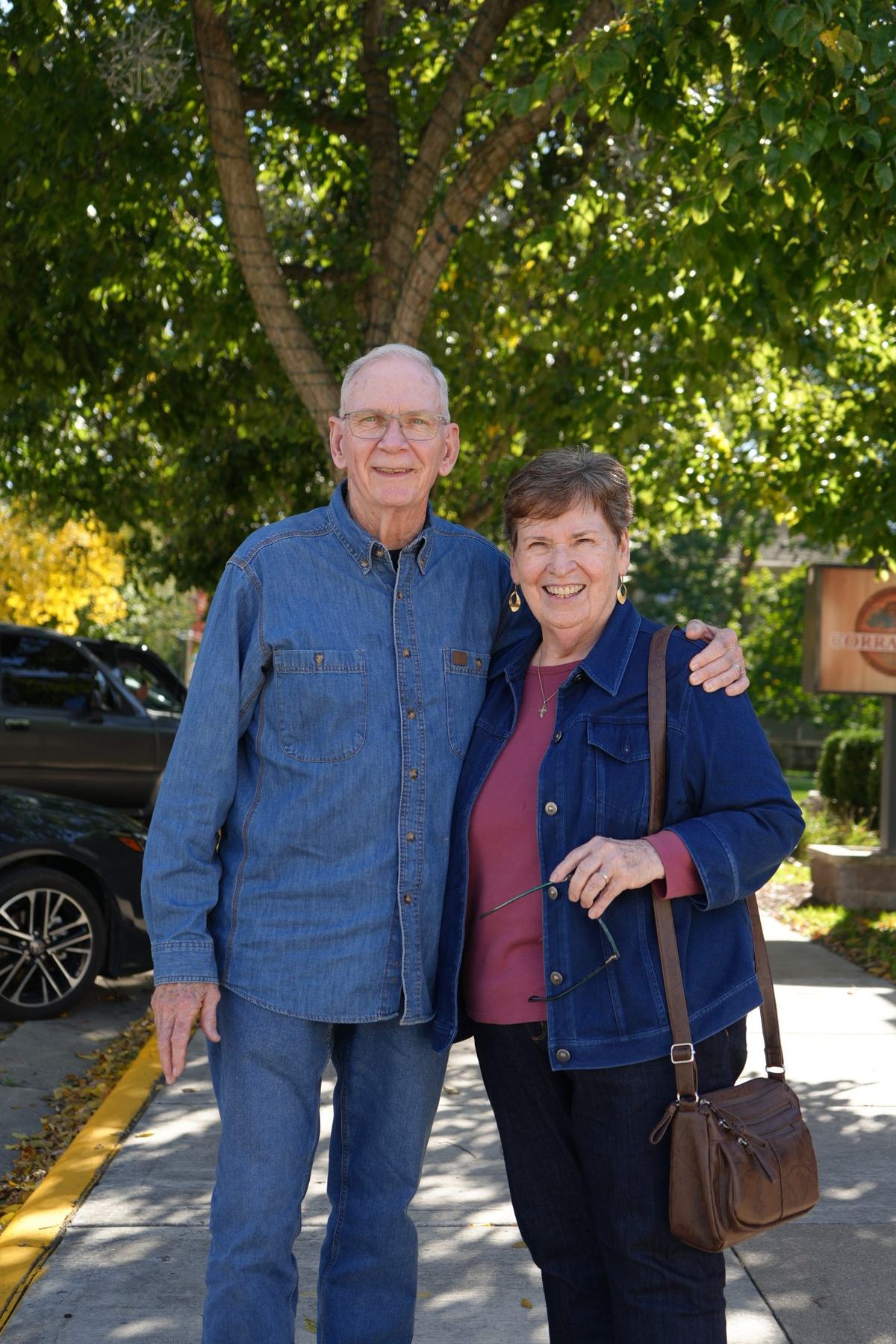 Two people wearing blue denim jackets smile at the camera beneath a leafy tree. One has an arm around the other's shoulders, and parked cars are visible behind them.