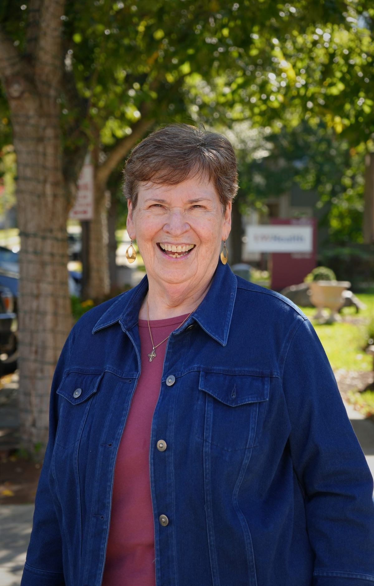 A person with short brown and gray hair wears a blue denim jacket, purple shirt, gold earrings and a cross necklace while smiling at the camera beneath a leafy tree.