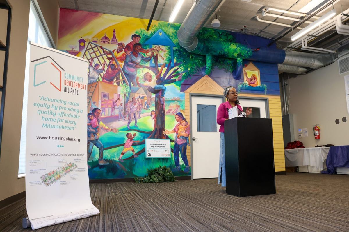 A person stands at a podium beside a Community Development Alliance banner, with a colorful mural depicting people and homes behind the person.
