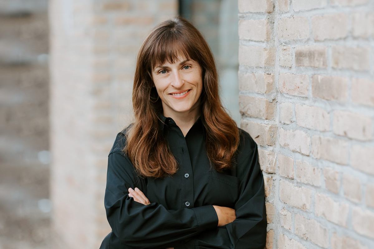 A person with long brown hair and bangs wears a black button-up shirt and stands with arms crossed beside a light brick wall.