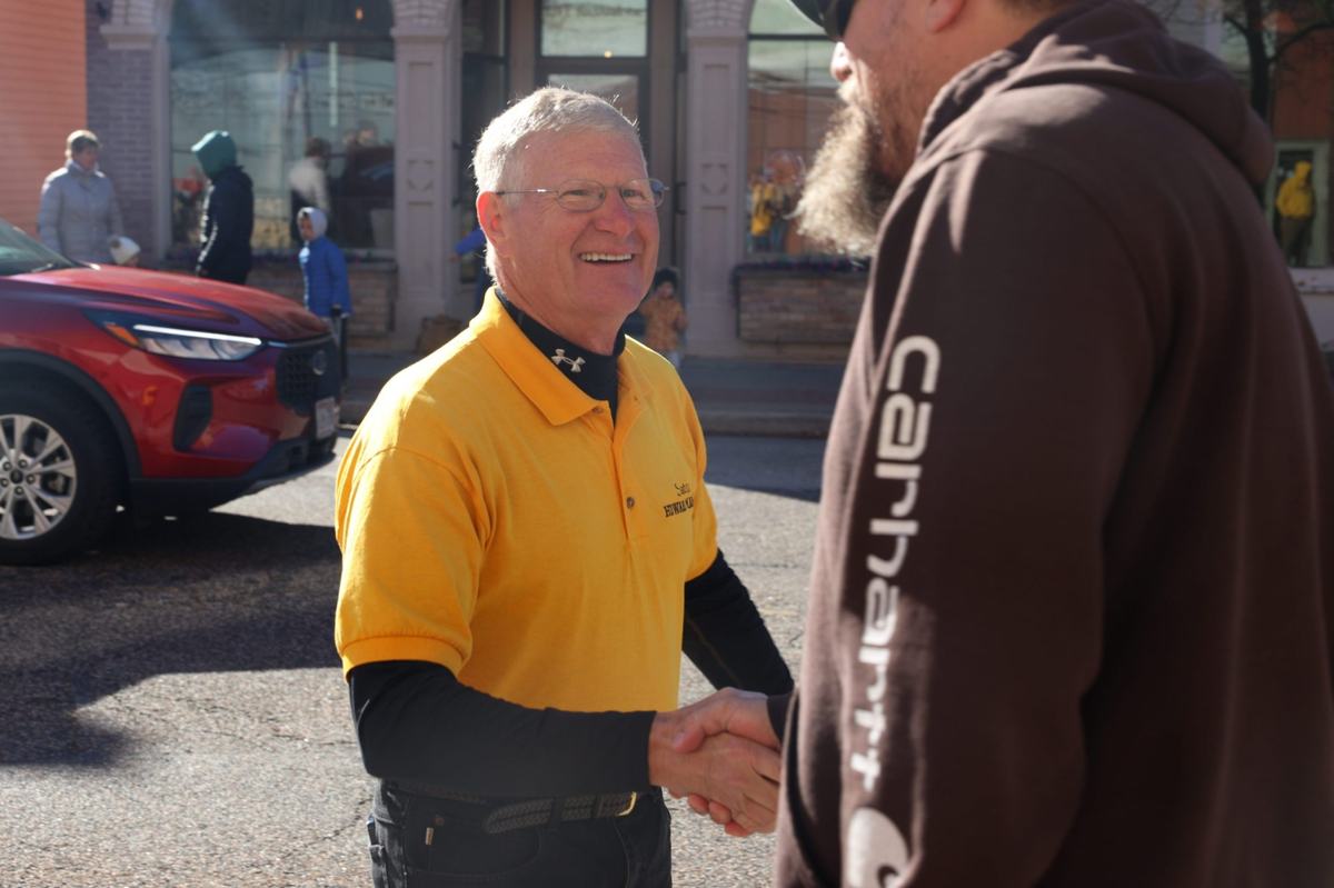 A person with gray hair and glasses wearing a yellow polo shirt smiles while shaking hands with a person in a brown hoodie on a street, with a red vehicle and pedestrians nearby.