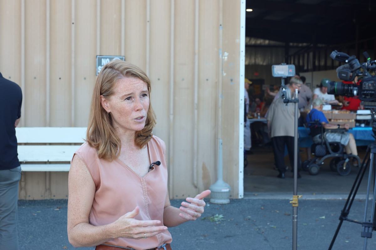 A person wearing a pink sleeveless blouse and a clip-on microphone gestures while standing near a video camera outside a building where people gather around tables.
