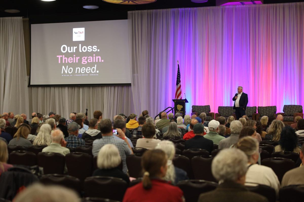 A person holds a microphone onstage before a seated audience. A projected image reads "Our loss. Their gain. No need."