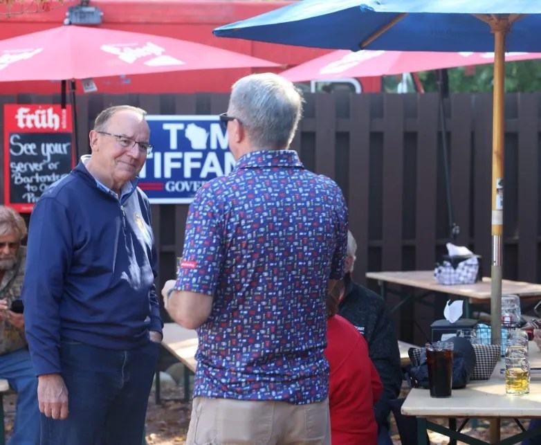 Two people stand facing each other near outdoor picnic tables and umbrellas, with a "Tom Tiffany Governor" campaign sign on a fence behind them.