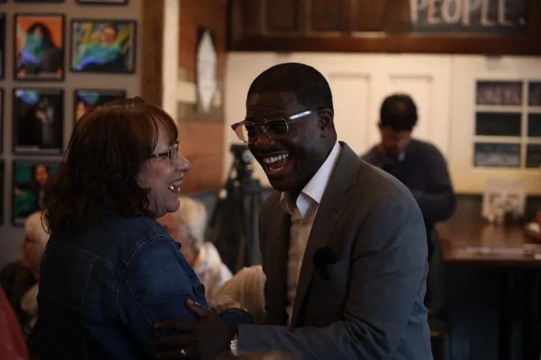Two people wearing glasses smile with their mouths open while facing each other. A person in a gray suit rests a hand on the arm of a person in a denim jacket.