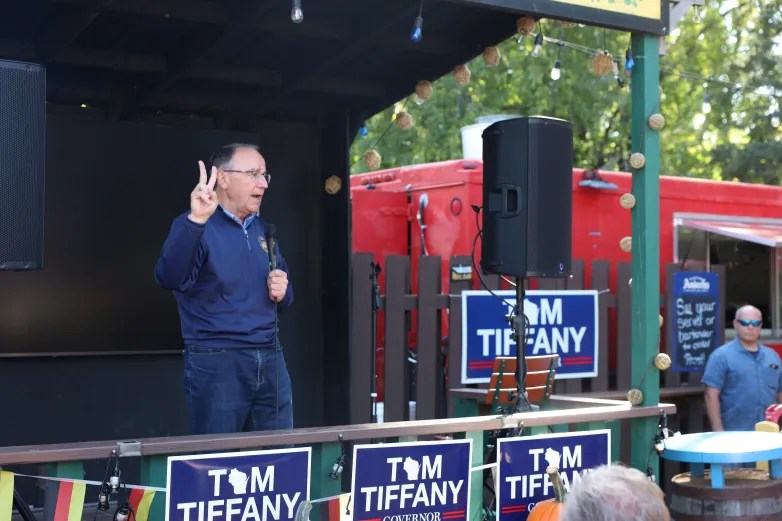 A person in a blue pullover holds a microphone and raises two fingers on an outdoor stage decorated with "Tom Tiffany Governor" campaign signs.