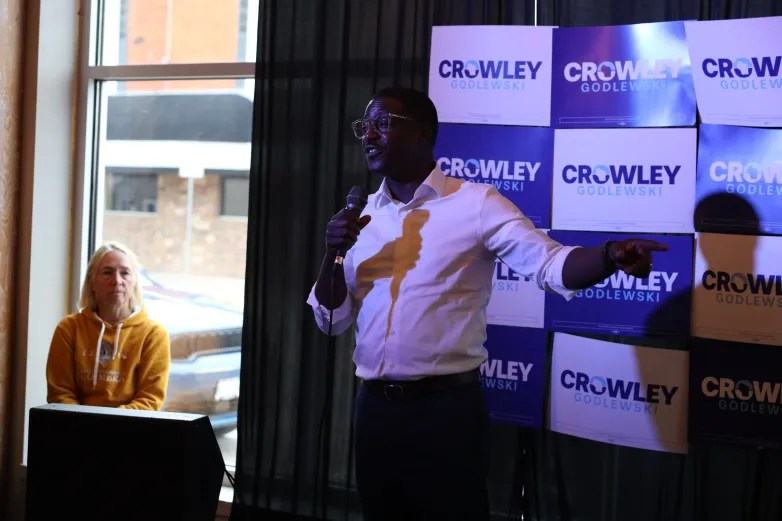 A person in a white shirt holds a microphone and points to the right in front of a wall of "Crowley Godlewski" signs. Another person sits nearby next to a window.