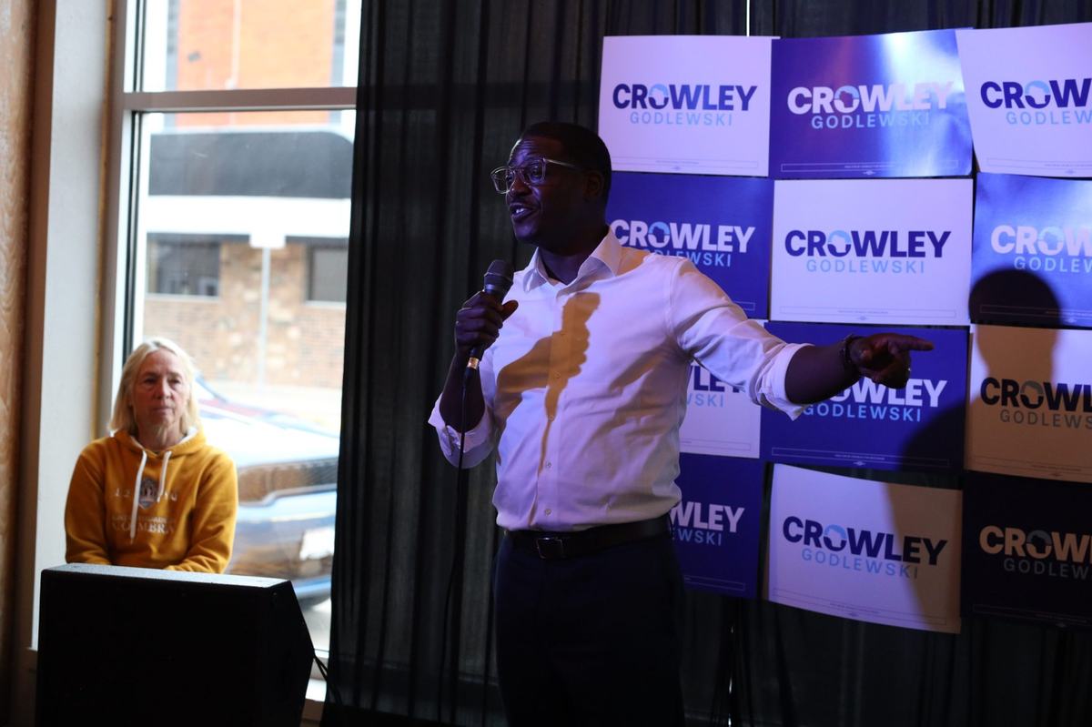 A person in a white shirt holds a microphone and points to the right in front of a wall of "Crowley Godlewski" signs. Another person sits nearby next to a window.