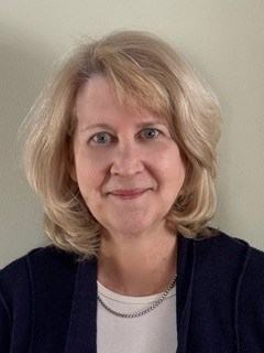 A person with shoulder-length blond hair wears a dark cardigan over a white shirt and a necklace against a light-colored wall.