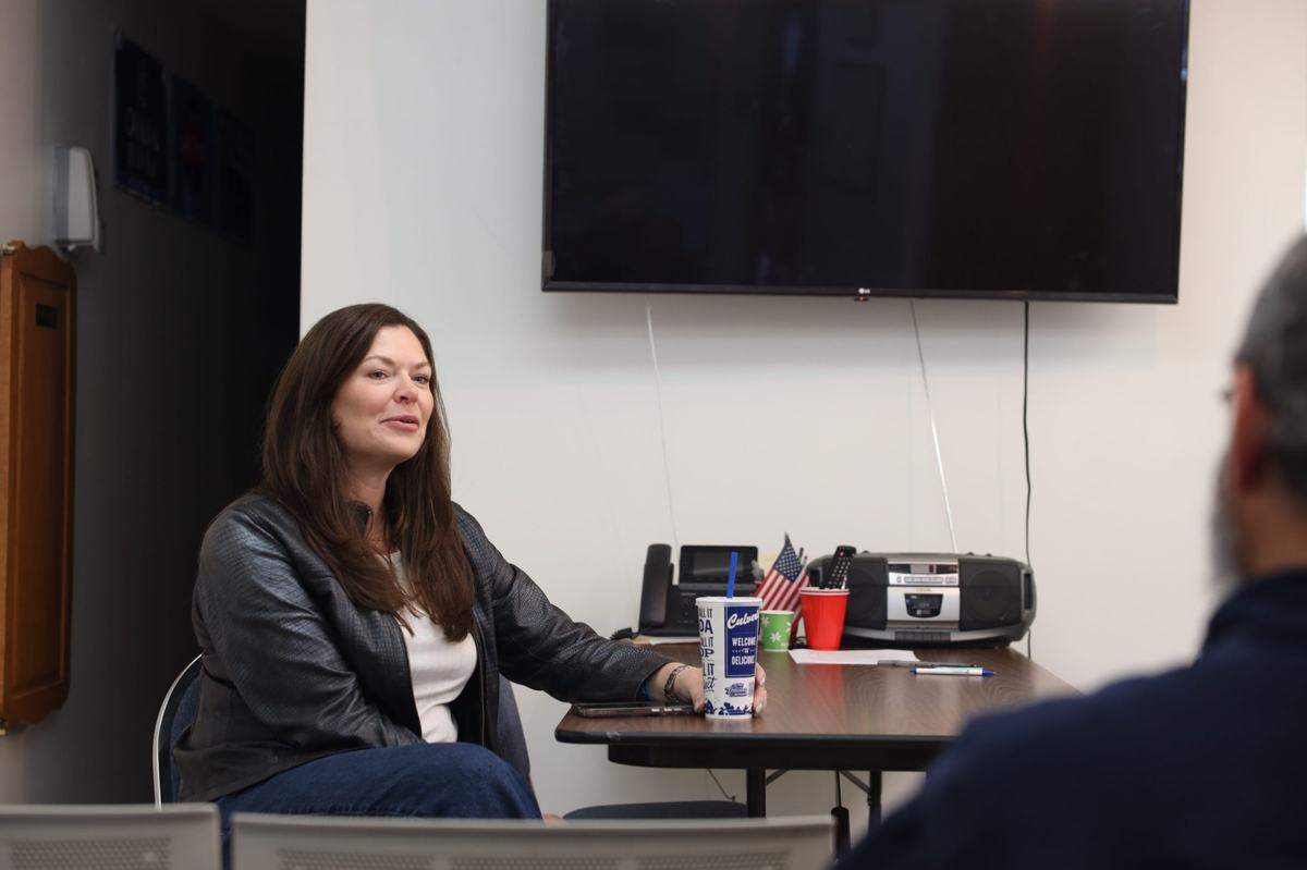 A person with long brown hair wearing a black jacket sits at a table holding a drink cup while speaking to another person. A television, radio and small American flag are nearby.