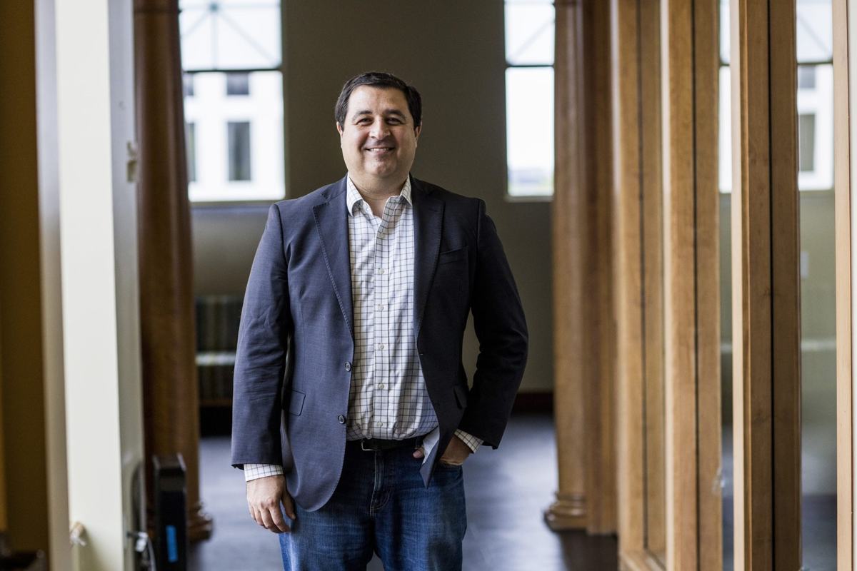A person in a dark jacket, checkered shirt and jeans stands with one hand in a pocket inside a room with tall windows and wooden columns.
