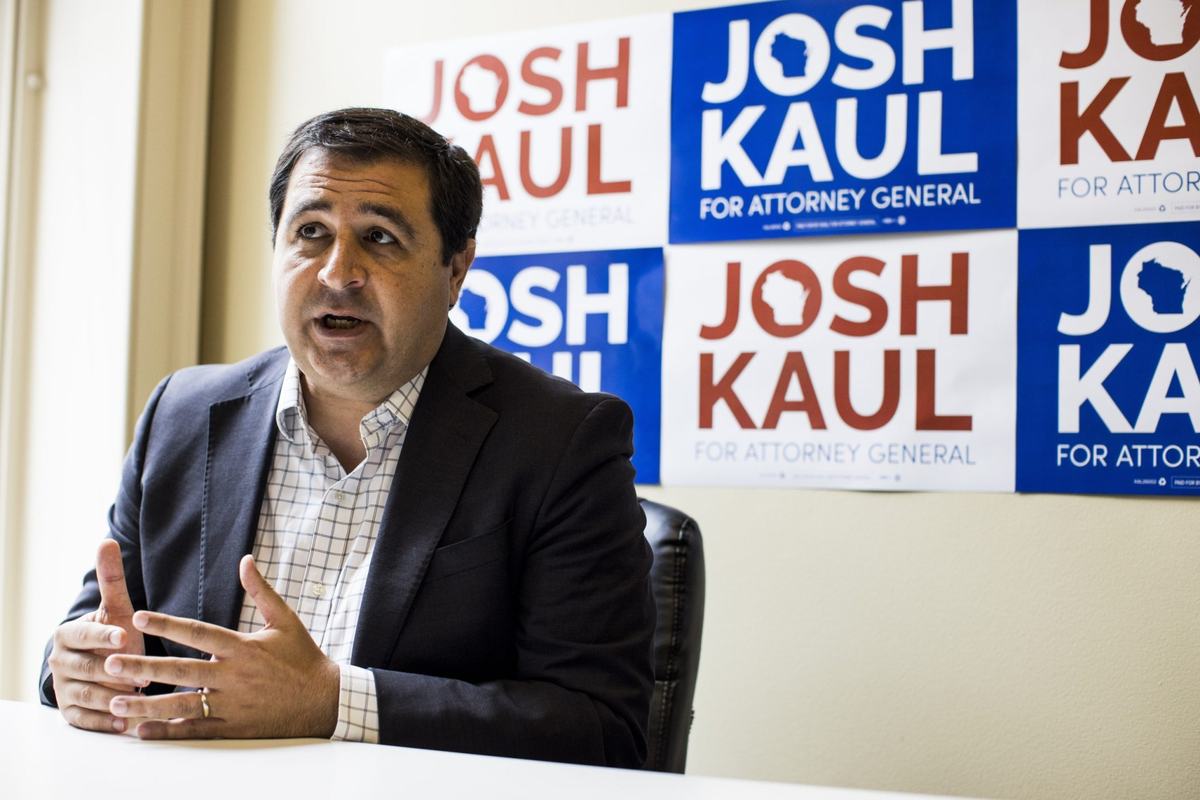 A person in a dark jacket gestures while seated at a table in front of signs reading "JOSH KAUL FOR ATTORNEY GENERAL"