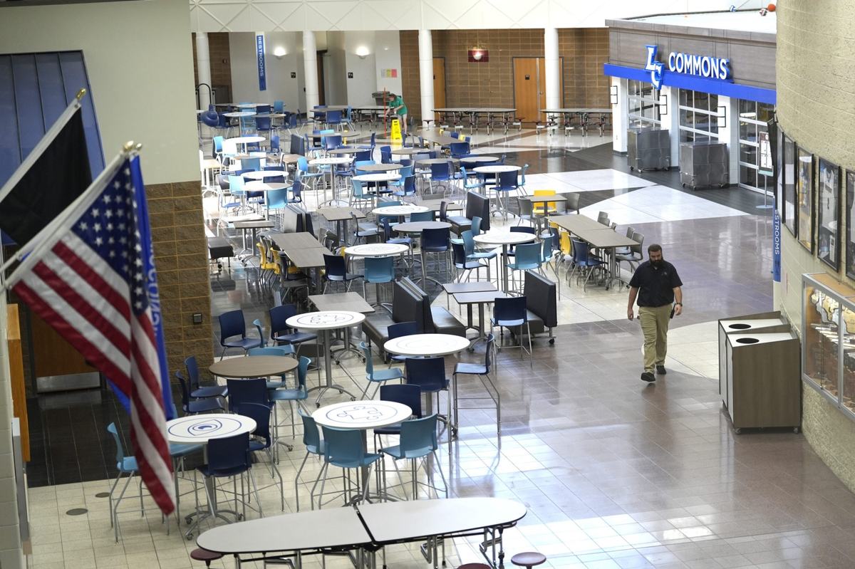 A person walks through a commons area filled with tables and chairs, with another person in the background, an American flag in the foreground and a sign reading "LC COMMONS" above doors.