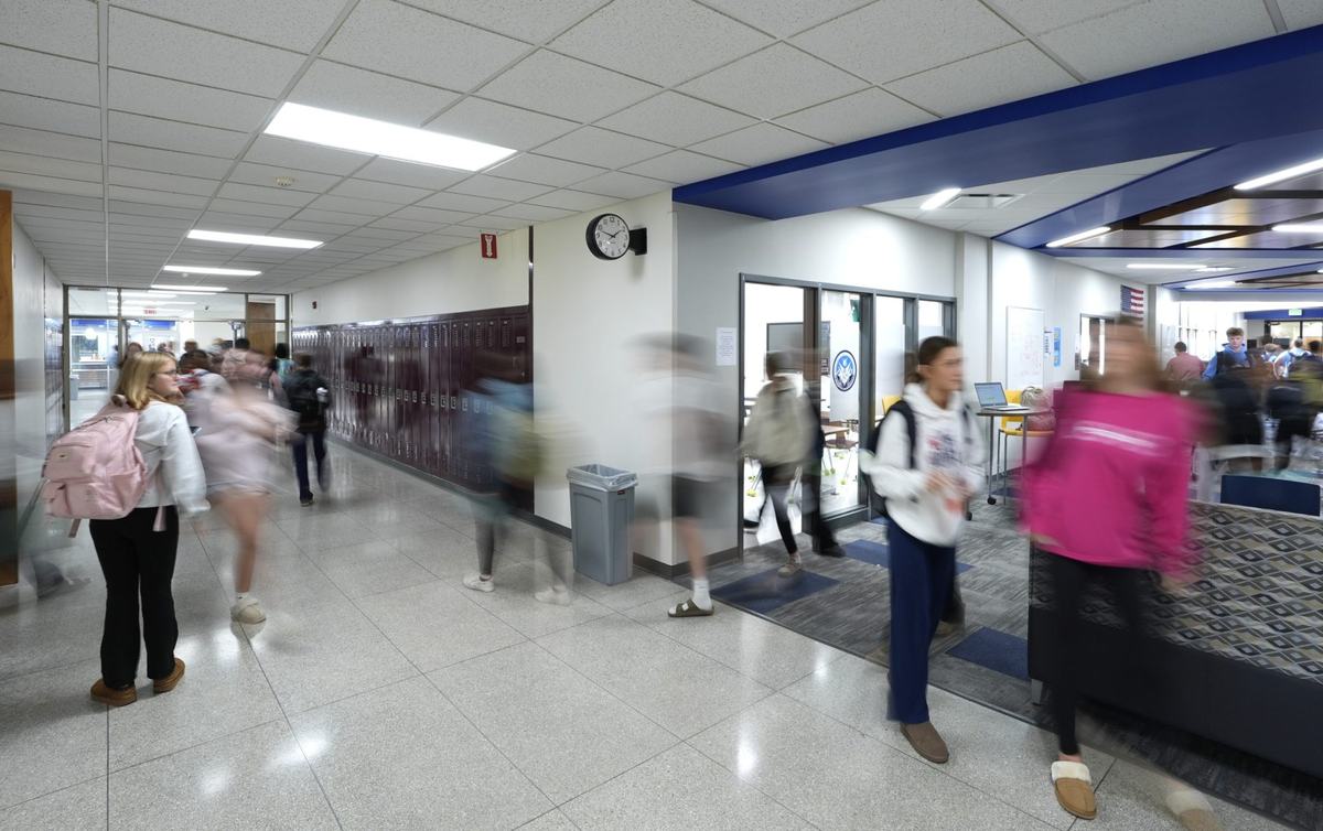 People carrying backpacks walk through a hallway lined with lockers, with several figures blurred by motion.