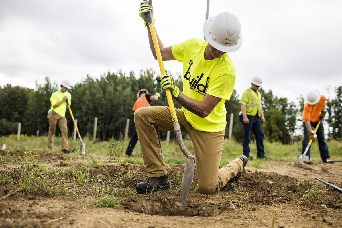A person wearing a white hard hat and neon yellow shirt reading "build my future" kneels and digs with a shovel in a dirt field as other people work with shovels in the background.